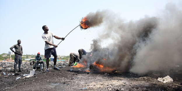 Mehrere Männer stehe auf einem Schrottplatz in Ghana und verbrennen alte Kabel und Elektroschrott. Die Luft ist voll mit grauem Dampf