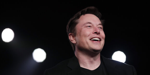 Tesla-Chef Elon Musk steht lachend im Scheinwerferlicht..