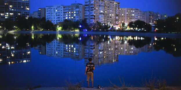 Ein Mann steht in der Dunkelheit an einem kleinen See vor Plattenbauten in der Stadt Bukarest und angelt