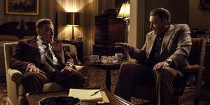 Zwei Männer im Anzug sitzen in einem schummerigen Zimmer in Sesseln.