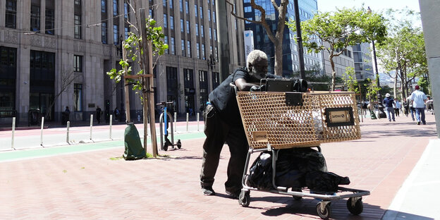 Ein schwarzer Mann schiebt einen Einkaufswagen mit seinen Sachen durch eine Straße mit eleganten Hochhäusern in San Francisco