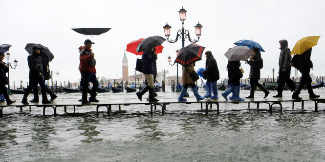 Menschen mit Schirmen laufen über einen Steg, der über das Wasser führt, dass auf dem Markusplatz steht