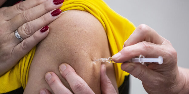 Eine Person bekommt eine Impfung in den Oberarm. Der Mensch trägt ein gelbes Oberteil und hat rote Fingernägel.