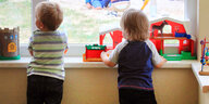 Zwei Kleinkinder stehen an einer Fensterbank und spielen mit Spielzeug.