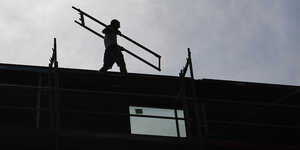 Ein Bauarbeiter trägt ein Element eines Baugerüstes über das Dach eines Rohbaus. Seine Silhouette zeichnet sich gegen den getrübten Himmel ab.