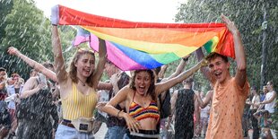 Drei junge Menschen halten im Regen einen Regenbogenfahne über ihre Köpfe.