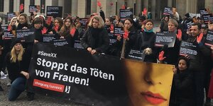 schwarz gekleidete Menschen vor dem Brandenburger Tor. Vor ihnen ein großes Plakat: Stoppt das Töten von Frauen an