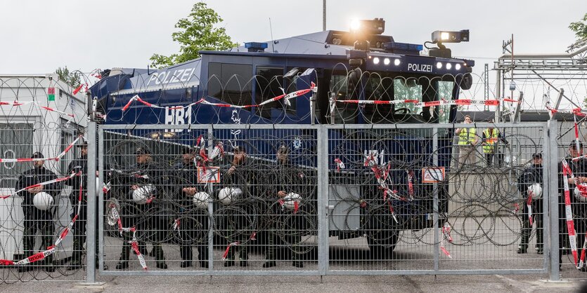 Hinter einem Zaun mit Stacheldraht stehen Polizisten und ein Wasserwerfer