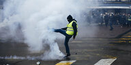 Eiine vermummte Person mit gelber Weste wirft einen Tränengasbehälter weg. Sie steht auf einer Straße in Paris. Andere Menschen im Hingtergrund beobachten die Szene.