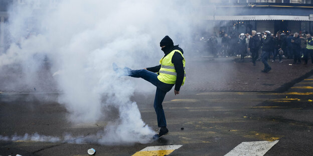 Eiine vermummte Person mit gelber Weste wirft einen Tränengasbehälter weg. Sie steht auf einer Straße in Paris. Andere Menschen im Hingtergrund beobachten die Szene.