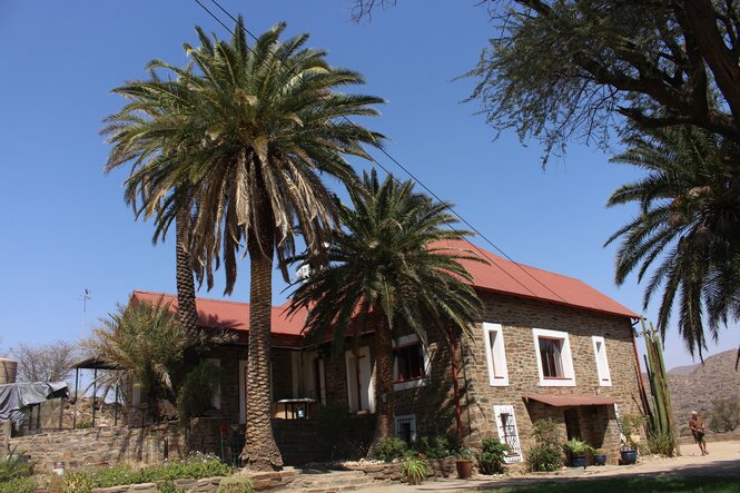 Ein Farmgebäude aus Steinen mit Spitzdach. Neben dem Haus steht ein hoher Kaktus. vor dem Haus stehen Palmen.