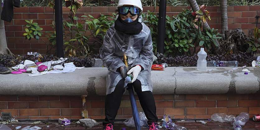 Eine junge Frau mit Schutzhelm und Atemmaske sitzt zwischen leeren Plastikflaschen auf einer Mauer