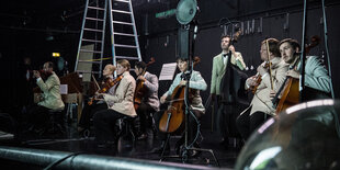 Musikerinnen und Musiker sitzen und stehen auf der Bühne im Residenztheater in München „Olympiapark in the Dark“ von Thom Luz. Sie spielen Geigen, Cellos und einen Bass.