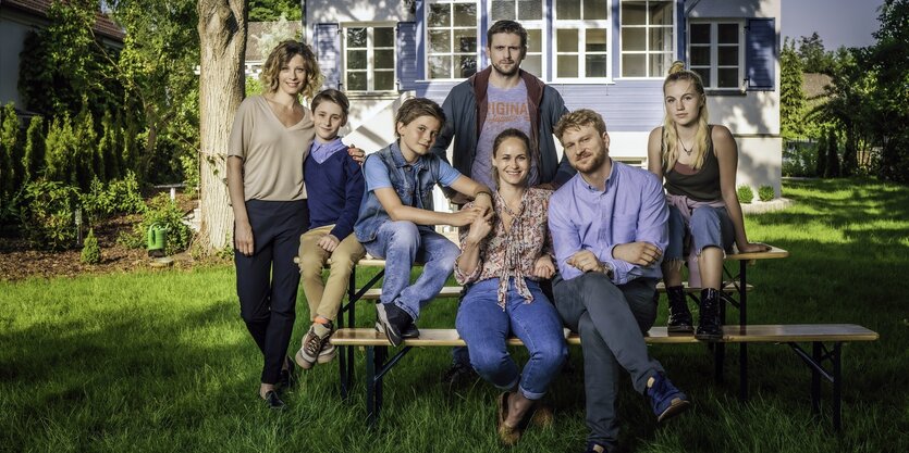 Junge Erwachsene und Kinder sitzen auf einer Bierbank vor einem Landhaus und lachen in die Kamera.