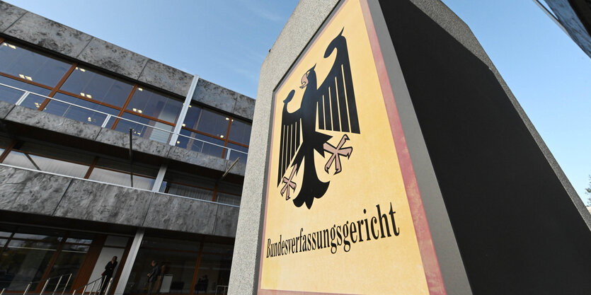 Außenansicht des Bundesverfassungsgerichtes. Eine Plakette auf einer Säule zeigt den Bundesadler. Im Hintergrund ist das gebäude selbst. Es sieht alt und heruntergekommen aus.