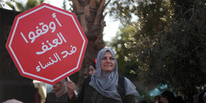 Eine Frau mit Kopftuch auf einer Demonstration. Sie hält ein Schild in die Luft, das wie ein Stopschild gestaltet ist, auf dem auf Arabisch steht: "Stoppt die Gewalt gegen Frauen!"