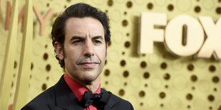 Sacha Baron Cohen, ein schlanker Mann mit dunklen kurzen Haaren steht vor einer goldenen Wand bei der Emmy-Verleihung 2019. Er trägt ein rotes Hemd, ein Jacket und eine Fliege.