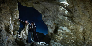 Durch den Eingang einer Höhle sieht man in den Nachthimmel, davor stehen ein Mann und eine Frau in flatternden Gewändern.