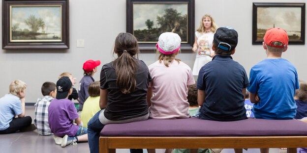 Kinder sitzen auf einer Bank und auf dem Boden vor Bildern in einem Museum