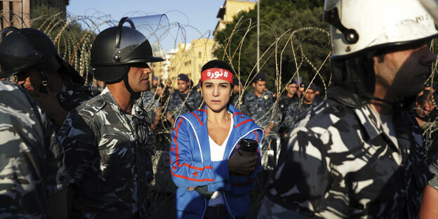 Eine Frau mit Stirnband (Aufschrift: Revolution) steht zwischen uniformierten Sicherheitskräften.