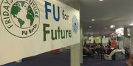 Ein Transparent mit der Aufschrift "FU for Future" und dem Logo von Fridays for Future hängt im Flur eines Gebäudes der FU. Daneben sieht man eine Sitzecke mit Sofas und Stühlen, umringt von Plakaten und Bannern. AktivistInnen sitzen auf den Sofas.