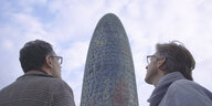 Zwei Männer stehen mit dem Rücken zum Betrachter vor einem zuckerhutförmigen Gebäude.