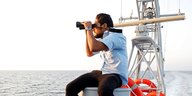 Ein Mann mit einem Fernglas sitzt neben Rettungsringen auf einem Schiff und schaut auf das Meer