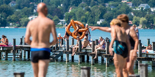 Menschen stehen in Badekleidung an einem Gewässer, in das eine Steg führt