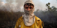 Ein Mann in gelber Feuerwejrkleidung und mit bedecktem Gesicht vor brennendem Wald