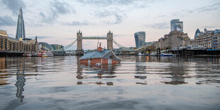 Die Atrappe eines typischen britischen roten Hauses sinkt in der Themse, im Hintergrund sieht man die Tower-Bridge