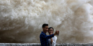 Ein junges Paar macht ein Selfie von sich, während im Hintergrund ein Wasserkraftwerk seine Tore öffnet.