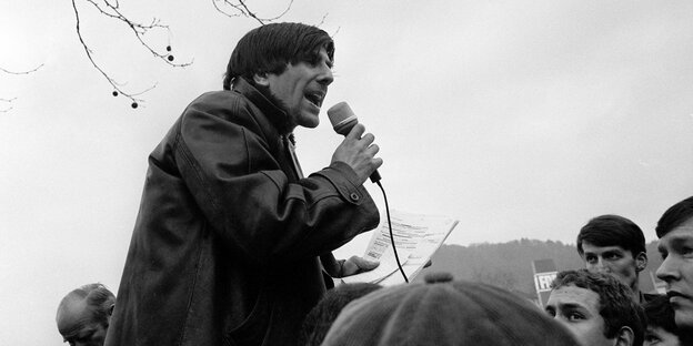 Auf dem schwarz-weiß-Bild sieht man einen Mann in Lederjacke, der vor einer Menschenmenge in ein Mikro spricht
