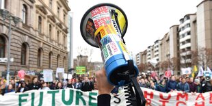 Eine Hand hält ein Megafon in die Luft, im Hintergrund sieht man Demonstrantinnen und Demonstranten mit einem Transparent