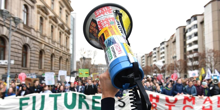 Eine Hand hält ein Megafon in die Luft, im Hintergrund sieht man Demonstrantinnen und Demonstranten mit einem Transparent