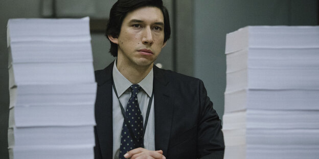 Ein Mann sitzt vor zwei hohen Papierstapeln.