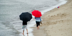 Ein schwarzer und ein roter Regenschirm mit Mensch drunter laufen am Strand nebeneinander
