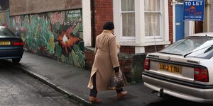 Eine Frau geht an einem Haus vorbei, auf dessen Wand eine Malerei ist, die dem Künstler Banksy zugeschrieben wird