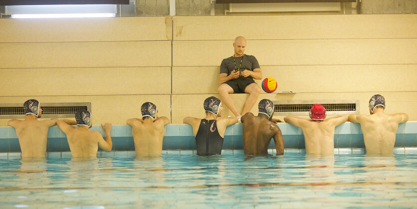 Ein Mann in Sportkleidung sitzt am Schwimmbeckenrand, vor ihm im Wasser sitzt eine Schwimmmannschaft.