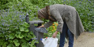 Frau im Mantel sucht in einer Mülltonne im Park nach Flaschen