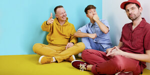 Drei Männer, jeder in einfarbiger Kleidung, sitzen im Schneidersitz auf dem Boden