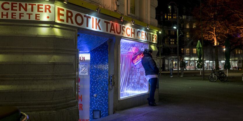 Eine Frau lehnt an einem lila beleuchteten Schaufenster und schaut auf einen dunklen Platz