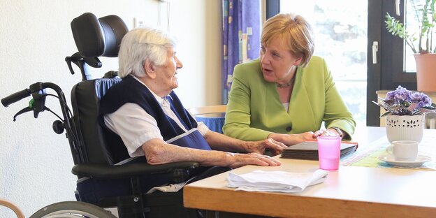 Eine Seniorin im Rollstuhl sitzt am Tisch mit Bundeskanzlerin Angela Merkel