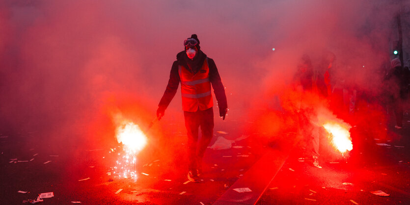 Ein Demonstrant in Paris zündet ein bengalisches Feuer. Man sieht noch eins neben ihm auf der Straße.