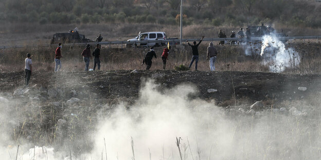 Menschen stehen im Westjordanland auf einem Hügel. Im Vordergrund schwebt Rauch. Die Menschen drehen sich zum Biuldhintergrund, wo hinter Leitplanken israelische Streitkräfte stehen.