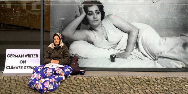 Die Autorin Stefanie de Velasco sitzt auf dem Boden mit einer Diddl-Maus Decke über den Beinen vor einen großen Fenster vor der Akademie der Künste in Berlin