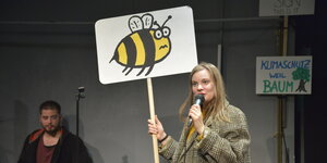 Ein Mädchen steht mit einem Plakat auf der Bühne. Auf dem Plakat ist eine Biene abgebildet. Im Hintergrund steht ein Plakat mit der Aufschrift "Klimaschutz weil Baum"