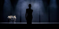 Im Vordergrund ist die Silhouette einer Figur zu sehen, hinten stehen viele Tänzerinnen und Tänzer eng zusammen.