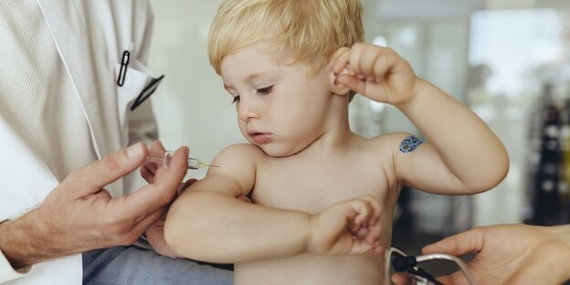 Ein Kind im Krabbelalter schaut auf seinen Arm, ein Arzt impft es gerade.