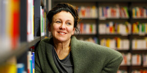 Olga Tokarczuk in einer Bibliothek, sie lehnt sich an ein Ragl an und lächelt wach in die Kamera