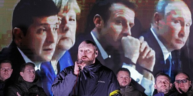 Männer stehen vor einer Videoleinwand auf der Selensky, Merkel, Macron und Putin zusehen sind